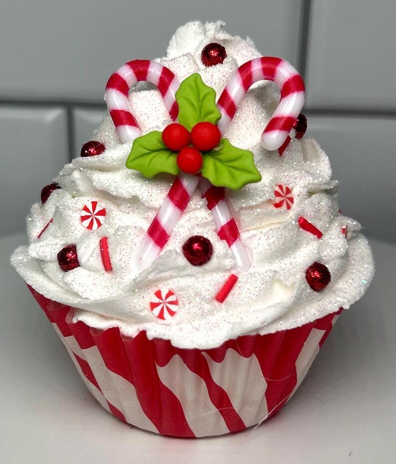 May include: A festive cupcake with white frosting, red and white candy canes, and holly berries. The cupcake is decorated with red sprinkles and candies, and sits in a red and white striped paper cup. A holiday-themed treat.