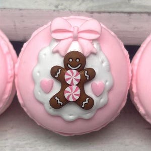 May include: Three pink macarons with gingerbread man decorations. Each macaron features a gingerbread man with a candy center, a pink bow, and heart accents on a white scalloped background.