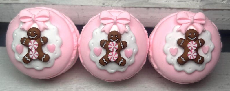 May include: Three pink macarons with gingerbread man decorations. Each macaron features a gingerbread man with a candy center, a pink bow, and heart accents on a white scalloped background.