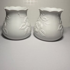 May include: Two white ceramic vases with a floral relief design. The vases have a ruffled top edge and a rounded body. The floral design includes flowers and leaves. The vases are displayed on a white surface.