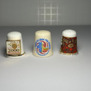 May include: Three ceramic thimbles with different designs. One thimble has "The New 2000 Millennium" text, another has "I Want to Take You Higher" text, and the third has "The Golden Jubilee" text with a red and gold pattern.