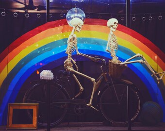 Pode incluir: Duas figuras de esqueleto andando de bicicleta em frente a um fundo de arco-íris. O esqueleto da frente está usando um chapéu e o esqueleto de trás está segurando o guidão.
