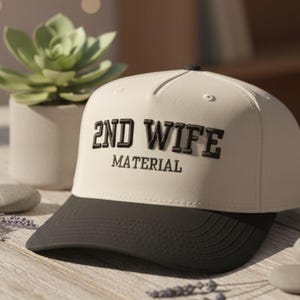 May include: A cream-colored baseball cap with a black brim. The cap features the embroidered text "2ND WIFE MATERIAL" in black block letters. The hat is a classic six-panel design.