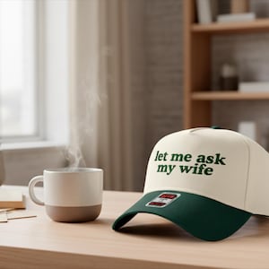 May include: A cream-colored baseball cap with a dark green brim and the text "let me ask my wife" in green. A white mug with a brown base sits next to the cap on a wooden table.