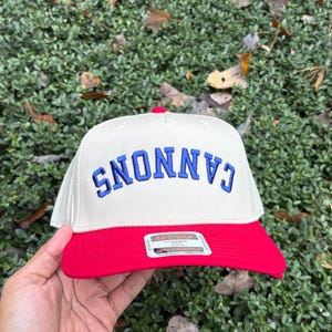 May include: A cream-colored baseball cap with a red brim and the word "CANNONS" embroidered in blue. The cap has a classic design and is likely made of a durable fabric. The background is a green bush.