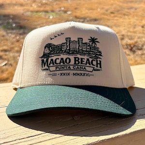 May include: A cream-colored baseball cap with a green brim. The cap features a green graphic of a building, palm tree, and the words "MACAO BEACH PUNTA CANA" in a stylized font. The brim is a solid dark green color.