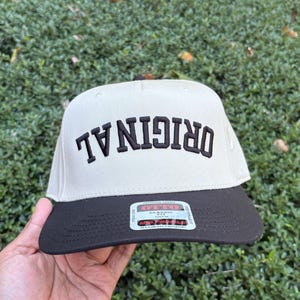 May include: A beige and black baseball cap with the word "ORIGINAL" embroidered in black. The cap has a flat brim and a structured fit. The cap is a classic style.