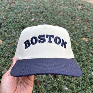 May include: A cream-colored baseball cap with a navy blue brim. The word "BOSTON" is embroidered in navy blue across the front of the cap. The cap is being held up by a hand.