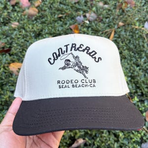 May include: A beige and black Contreras Rodeo Club hat. The hat features the text "Contreras" above a graphic of a cowboy riding a horse, and the text "Rodeo Club Seal Beach CA". The hat has a flat brim.