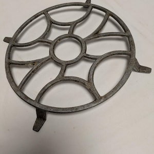Peut inclure: Un dessous-de-plat circulaire en fonte avec un motif ajouré décoratif. Il présente un anneau central et des segments incurvés, soutenus par trois petits pieds. Le métal a un aspect gris vieilli.
