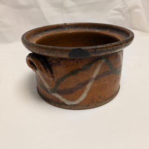 Pode incluir: Um pote de cerâmica rústico feito à mão com uma borda larga marrom escuro e duas pequenas alças. O pote apresenta um exterior marrom avermelhado texturizado com linhas abstratas pintadas em branco e marrom escuro. O interior é marrom mais escuro.