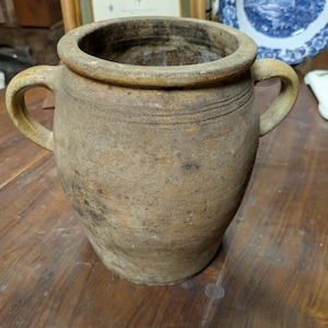 Może przedstawiać: Rustykalny, antyczny dzbanek ceramiczny z dwoma uchwytami. Dzbanek ma szeroki otwór i grubą, zaokrągloną krawędź. Zewnętrzna strona jest nakrapiana na brązowo z ciemniejszymi plamami, co sugeruje wiek i użytkowanie. Dzbanek stoi na drewnianej powierzchni.