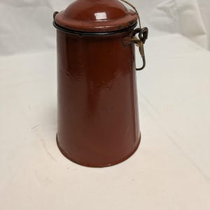Peut inclure: Un récipient en émail marron, cylindrique et vintage, avec un couvercle et une poignée. Le récipient a une finition brillante et un bord noir. Une ficelle est attachée au couvercle. Le récipient est probablement utilisé pour le stockage ou la cuisine.