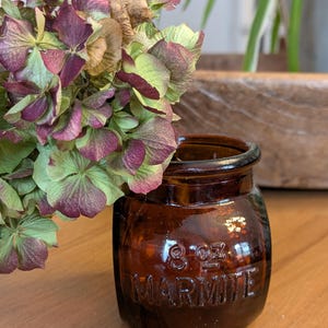 Peut inclure: Un petit pot en verre brun avec le mot "MARMITE" en relief sur le devant, ainsi que "8 oz.". Le pot est rempli d'une composition florale séchée de pétales verts, violets et bruns. Le pot est posé sur une surface en bois.
