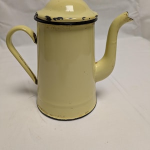 Peut inclure: Une cafetière en émail de couleur crème avec un couvercle et un bec courbé. La cafetière a un bord et une poignée noirs, montrant des signes d'usure. La conception comprend une poignée incurvée et un petit bouton sur le couvercle.