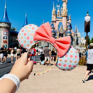 May include: A pair of mouse ears with a large coral bow and patterned ear details. The ears feature a geometric design in pastel colors with gold accents. The headband is held up in front of a castle.
