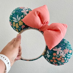 May include: Mouse ear headband featuring a coral bow and floral-patterned ears. The ears are covered in a dark teal fabric with a floral print in pink, yellow, and white. The headband is held by a hand.