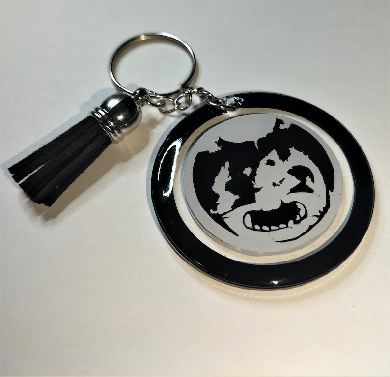 May include: A round black and white keychain featuring a cartoon character design. The keychain has a black border, a silver key ring, and a black tassel. The design is a black and white image of a cartoon character.