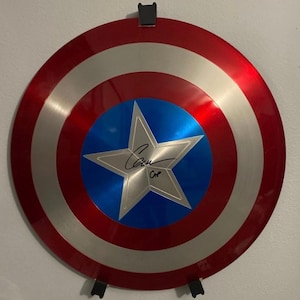 Puede incluir: Un escudo de Capitán América rojo, blanco y azul con una estrella plateada en el centro. El escudo está firmado "Chris Evans" con "Cap" debajo de la firma.