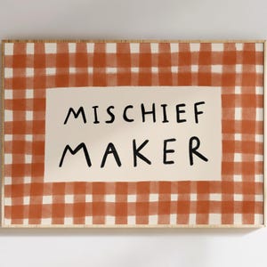May include: A framed print with a red and white gingham pattern. The print has the words "MISCHIEF MAKER" in black, bold letters on a cream-coloured rectangular background. The frame is a light brown colour.