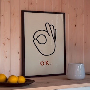 May include: A white kitchen with white cabinets and a wood wall. A framed print with a black and white illustration of a hand making the OK sign with the text "OK." is hanging on the wall. There are three lemons in a bowl on the counter.