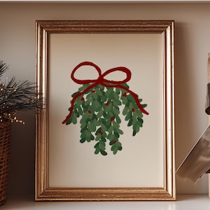 May include: A framed print of mistletoe with a red bow, displayed on a white shelf. A woven basket with greenery, decorative white houses, and a wrapped gift box with a brown ribbon are also on the shelf.