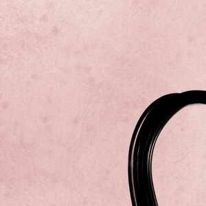 May include: A textured, light pink background surface featuring a partial, thick, glossy black circular frame or border element positioned on the right side, creating a stark contrast between the soft pastel color and the bold, dark shape.