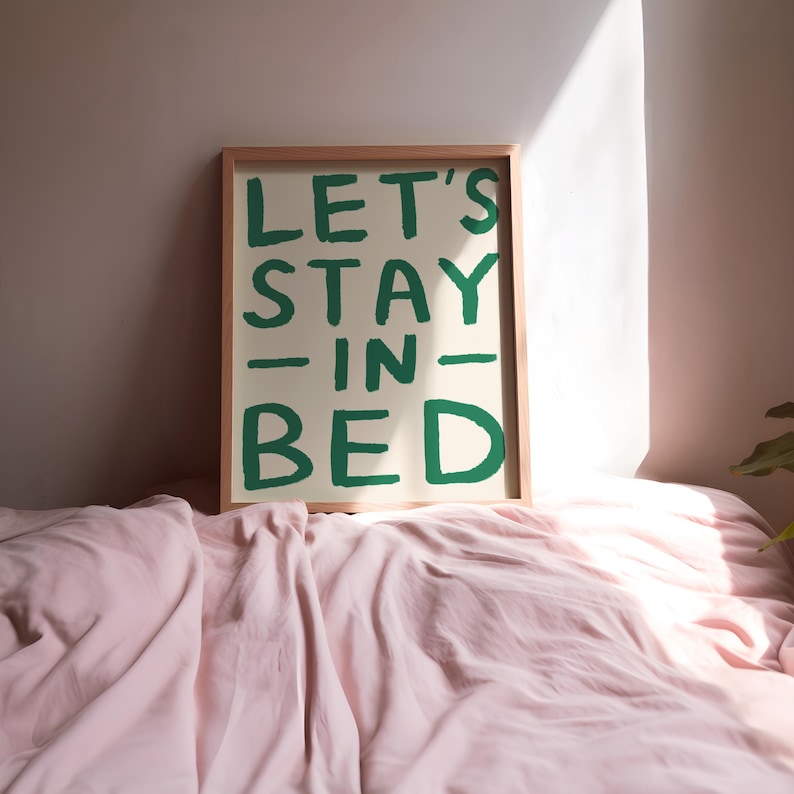 May include: A framed print with the text "LET'S STAY -IN- BED" in green lettering on a cream background. The print is sitting on a pink bedspread.