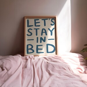 May include: A framed print with the text "LET'S STAY -IN- BED" in blue lettering on a cream background. The print is sitting on a pink bedspread.