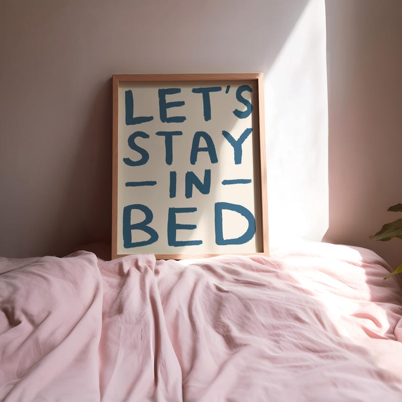 May include: A framed print with the text "LET'S STAY -IN- BED" in blue lettering on a cream background. The print is sitting on a pink bedspread.