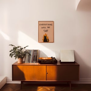 May include: A black cat sitting in flames with the text "Everything will be fine" on a peach background. The print is framed and hanging on a white wall above a mid-century modern wooden cabinet with a record player and a stack of vinyl records.