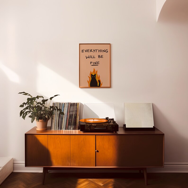 May include: A black cat sitting in flames with the text "Everything will be fine" on a peach background. The print is framed and hanging on a white wall above a mid-century modern wooden cabinet with a record player and a stack of vinyl records.