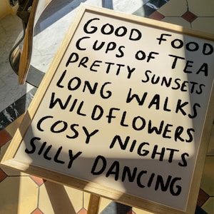 May include: A framed sign with the text "Good Food, Cups of Tea, Pretty Sunsets, Long Walks, Wildflowers, Cosy Nights, Silly Dancing" written in black marker on a beige background.
