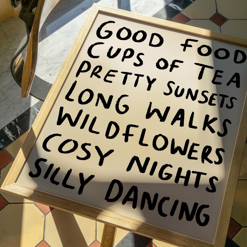 May include: A framed sign with the text "Good Food, Cups of Tea, Pretty Sunsets, Long Walks, Wildflowers, Cosy Nights, Silly Dancing" written in black marker on a beige background.