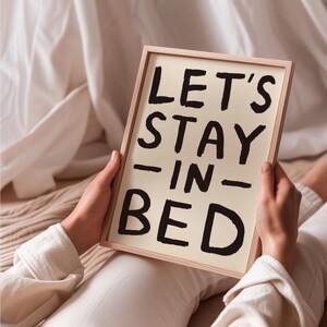 May include: A white sign with black lettering that reads "Let's Stay In Bed". The sign is being held by a person.