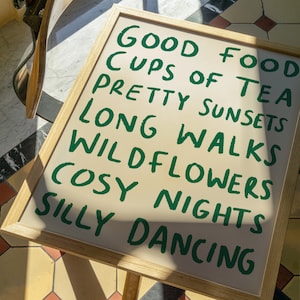May include: A framed print with the text "Good Food, Cups of Tea, Pretty Sunsets, Long Walks, Wildflowers, Cosy Nights, Silly Dancing" written in green on a beige background.