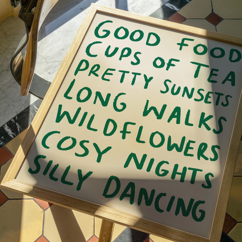 May include: A framed print with the text "Good Food, Cups of Tea, Pretty Sunsets, Long Walks, Wildflowers, Cosy Nights, Silly Dancing" written in green on a beige background.