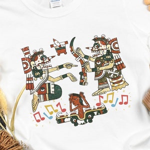May include: White t-shirt with a colorful graphic print of three figures playing music. The figures are stylized and have a traditional Aztec design. The graphic includes musical notes.