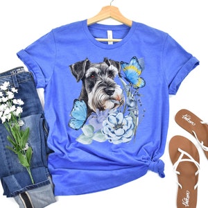 Può includere: Una maglietta blu reale con un'illustrazione ad acquerello di un cane schnauzer nano con farfalle blu e fiori.