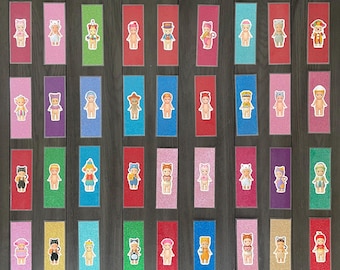 May include: A selection of glittery bookmarks in various colours, each with a sticker of a small doll in different outfits. The bookmarks are displayed on a dark wood surface. Colours include red, pink, blue, green, and yellow.