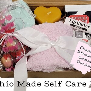 Könnte beinhalten: Eine braune Pappbox gefüllt mit Selbstpflegeartikeln. Die Box enthält ein rosa Handtuch, eine gelbe herzförmige Kerze, eine kleine Tüte mit rosa herzförmigen Süßigkeiten, eine kleine Dose Lippenbutter und eine Seife. Die Box ist mit einem weißen Band gebunden und hat einen rosa Zettel mit der Aufschrift "Wählen Sie aus 18 personalisierten Karten und Anlässen!" Die Box ist mit "Ohio Made Self Care Kit" beschriftet.