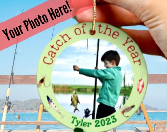 May include: A green and white round ornament with the text "Catch of the Year" and a photo of a boy fishing. The ornament has a gold string for hanging and the text "Tyler 2023" at the bottom.