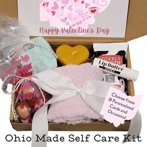 Può includere: Una scatola di cartone marrone con un design a forma di cuore rosa e bianco sul coperchio. La scatola è aperta e contiene una varietà di articoli per la cura di sé, tra cui un asciugamano da bagno rosa, una candela a forma di cuore, un piccolo sacchetto di bombe da bagno, un tubo di balsamo per le labbra e un piccolo sacchetto di sali da bagno. La scatola è etichettata "Ohio Made Self Care Kit" e "Scegli tra 18 carte personalizzate e occasioni!"