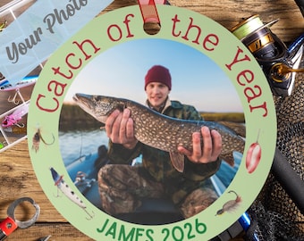 May include: A round, light green personalized ornament featuring a central photograph of a person holding a large fish. The text Catch of the Year is printed in red along the top, and the name JAMES 2026 is printed in green at the bottom.