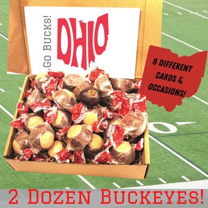 May include: A cardboard box filled with 2 dozen chocolate buckeyes, a type of peanut butter candy. The box is open and the buckeyes are visible. The box is decorated with the words "Go Bucks!" and "Ohio" in red letters. The box is sitting on a green and white football field. The text "8 DIFFERENT CARDS & OCCASIONS!" is visible on a red Ohio-shaped graphic. The text "2 DOZEN BUCKEYES!" is visible at the bottom of the image.