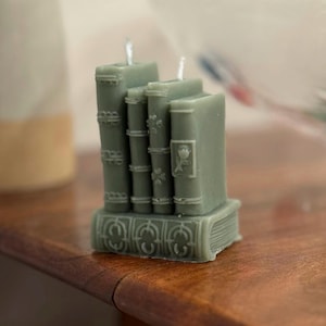 Puede incluir: Una vela verde salvia con forma de pila de libros. La vela presenta detalles de lomos de libros y una base decorativa. Las mechas son visibles en la parte superior. La vela está colocada sobre una superficie de madera.