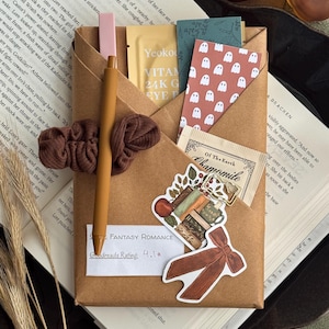 May include: A brown paper-wrapped book package featuring a brown scrunchie, a tan pen, a tea bag, a gold-toned paperclip, and stickers of ghosts, books, and a bow. A white card displays the text Fantasy Romance and Goodreads Rating: 4.1.