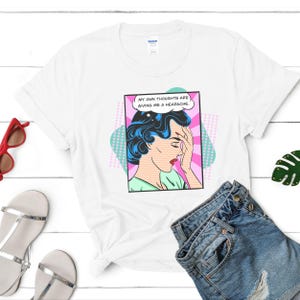 K&ouml;nnte beinhalten: Wei&szlig;es T-Shirt mit einer Grafik im Pop-Art-Stil einer Frau mit blauen Haaren, die sich an den Kopf fasst, und einer Sprechblase mit dem Text "Meine eigenen Gedanken bereiten mir Kopfschmerzen."