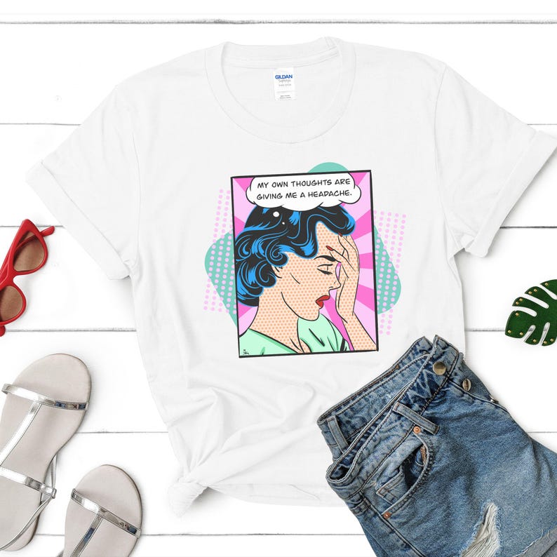 K&ouml;nnte beinhalten: Wei&szlig;es T-Shirt mit einer Grafik im Pop-Art-Stil einer Frau mit blauen Haaren, die sich an den Kopf fasst, und einer Sprechblase mit dem Text "Meine eigenen Gedanken bereiten mir Kopfschmerzen."