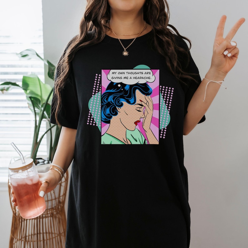 K&ouml;nnte beinhalten: Schwarzes T-Shirt mit einer Pop-Art-Grafik einer Frau mit Kopfschmerzen. Die Grafik enth&auml;lt eine Sprechblase mit dem Text "Meine eigenen Gedanken bereiten mir Kopfschmerzen."